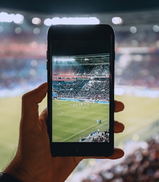 A hand holding a smartphone captures a live view of a soccer stadium filled with fans and players on the field. The image on the smartphone screen is crisp, showing a wide-angle view of the game in action.